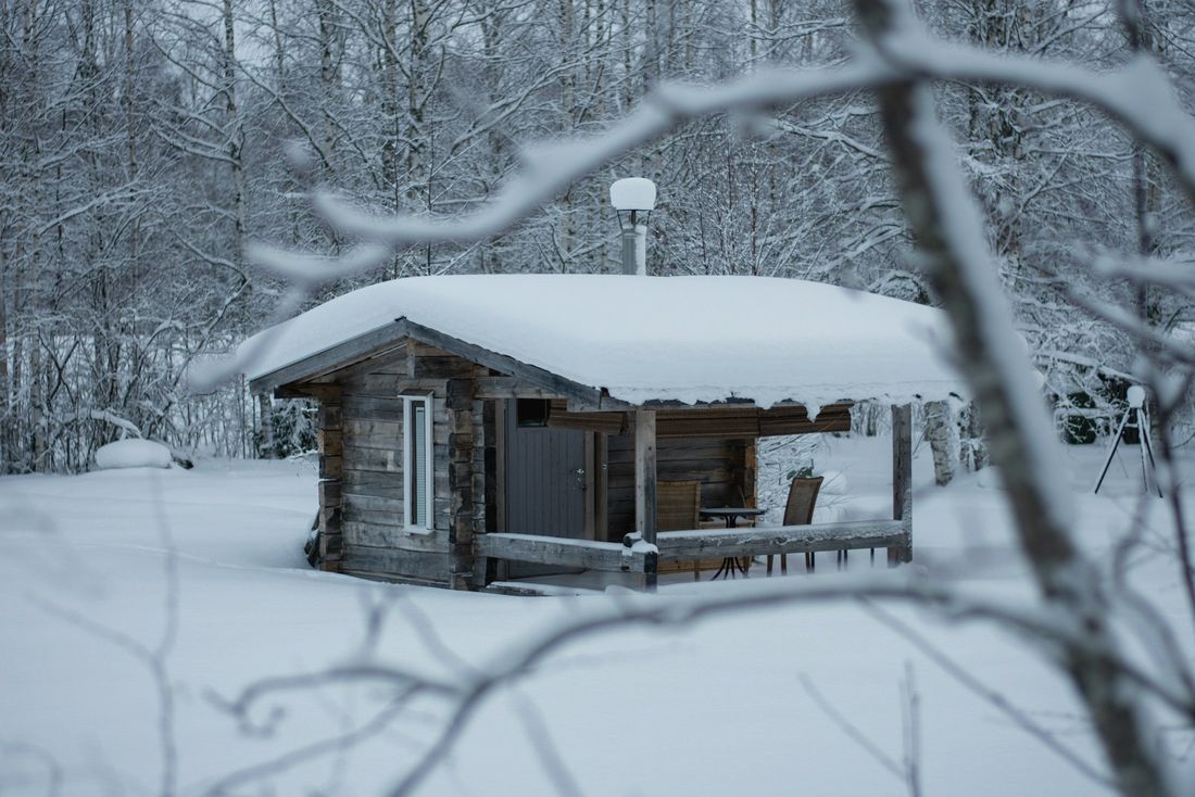 Pieni hirsisauna lumisessa metsässä. Katolla on paksu lumikerros.