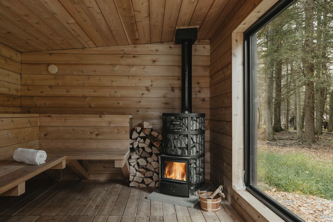 Puupaneloitu löylyhuone, jossa on puulämmitteinen kiuas, halkopino ja leveä ikkuna metsään.