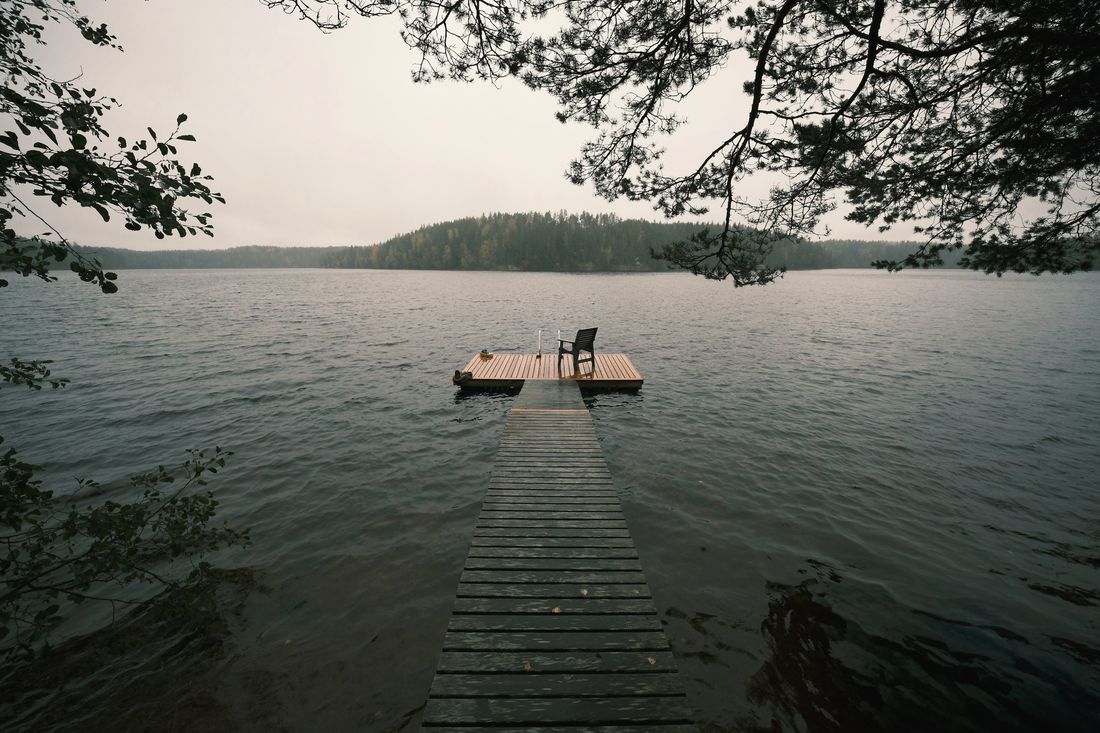 Kapea puulaituri johtaa tyynellä järvellä kelluvalle uintilaiturille, jolla on tuoli.