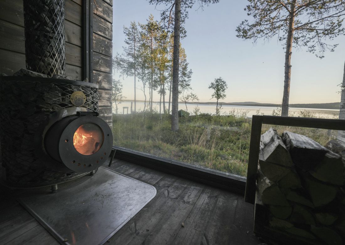 Musta kamiina peltilevyn päällä ja lattiaan asti ulottuva ikkuna. Ikkunasta näkyy koivuja, mäntyjä ja järvi.