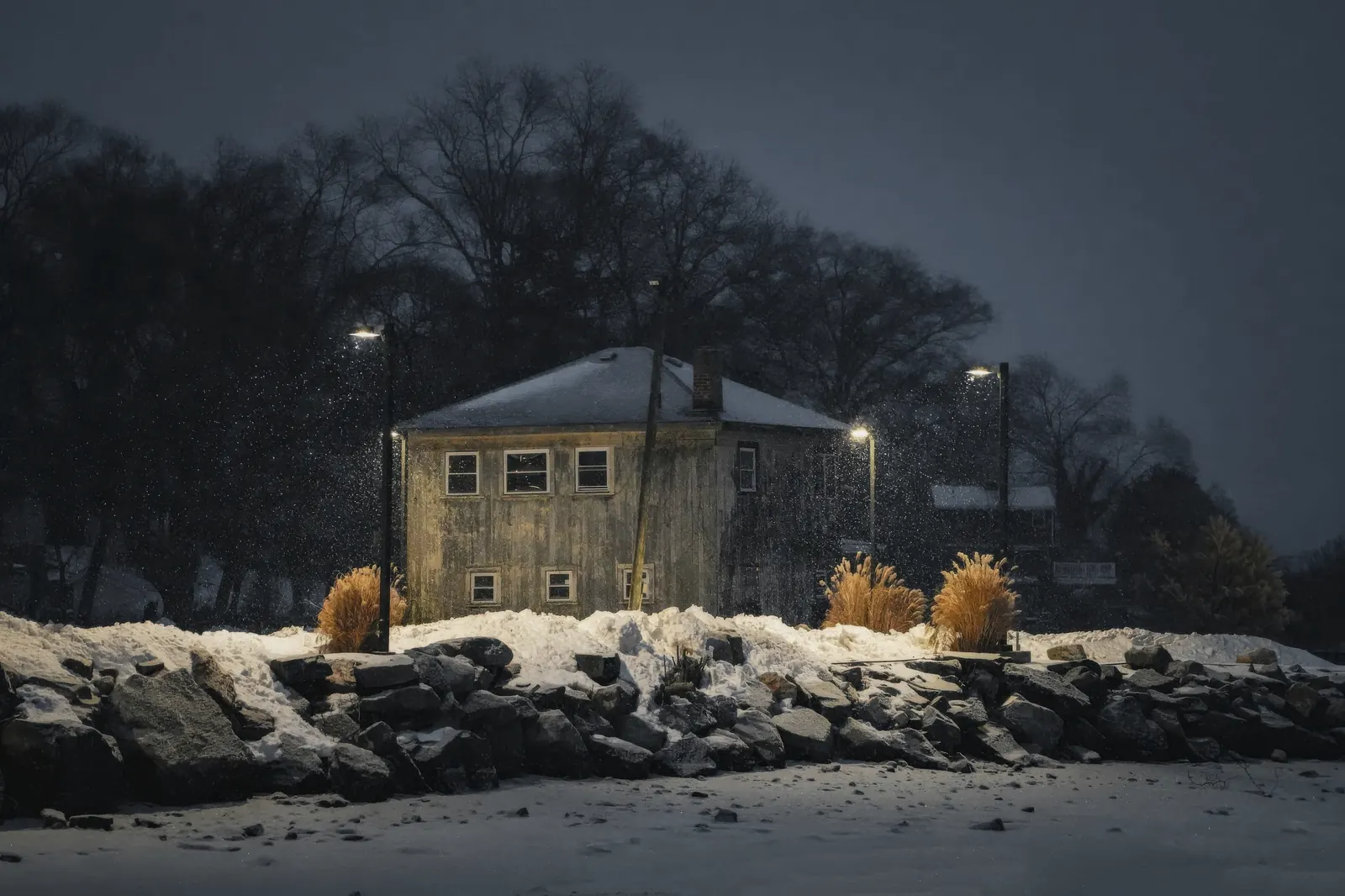Puutalo rannalla talviyönä, ikkunoissa valoa ja lunta kallioilla