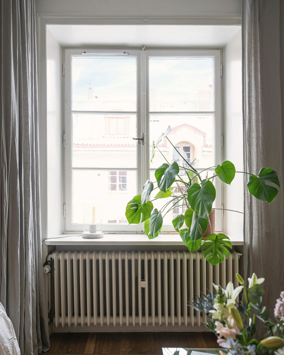 Korkea ikkuna, valurautapatteri ja harmaat pellavaverhot. Ikkunalaudalla on peikonlehti.