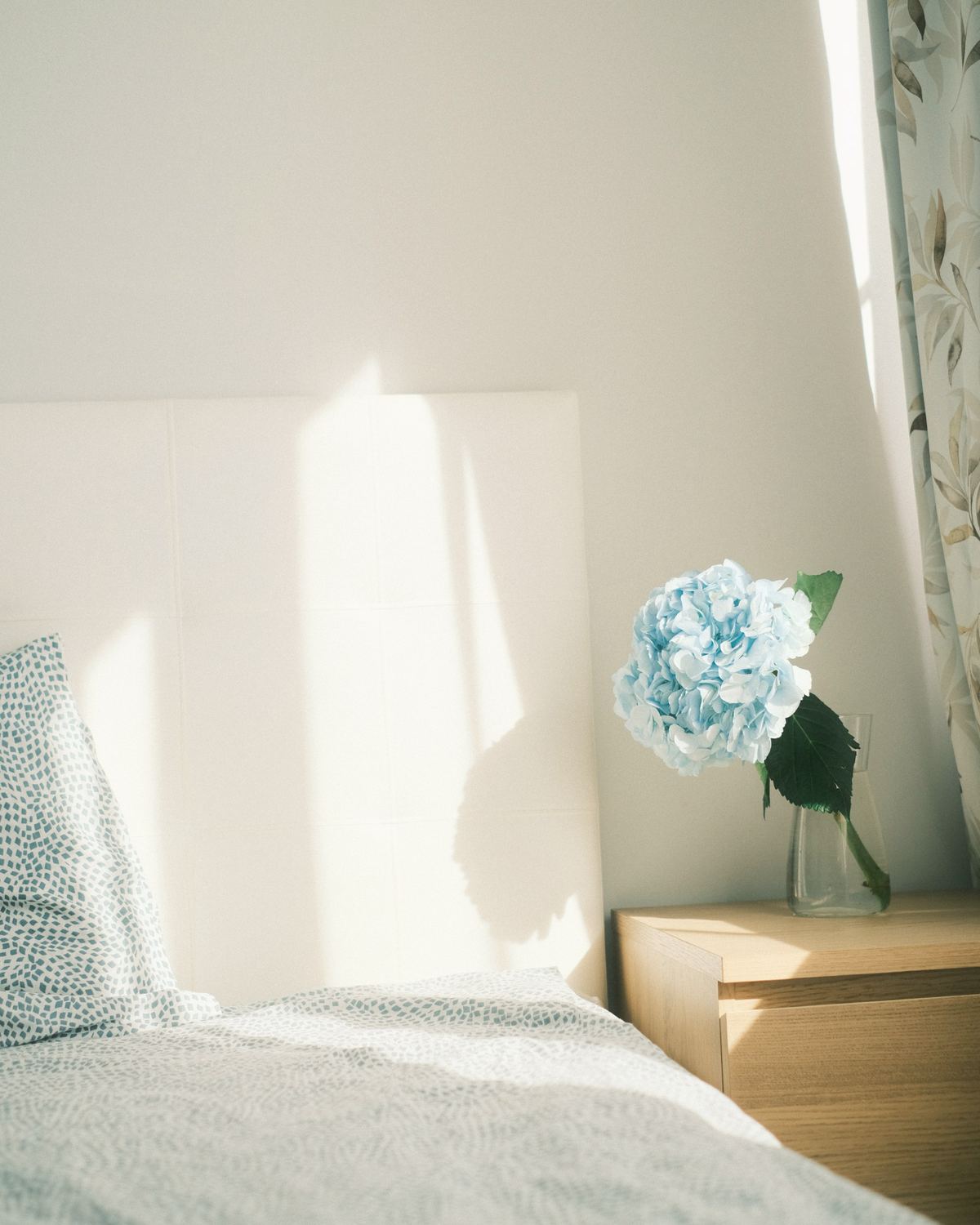 Makuuhuoneen sänky ja vaalea yöpöytä, jolla on hortensia maljakossa.