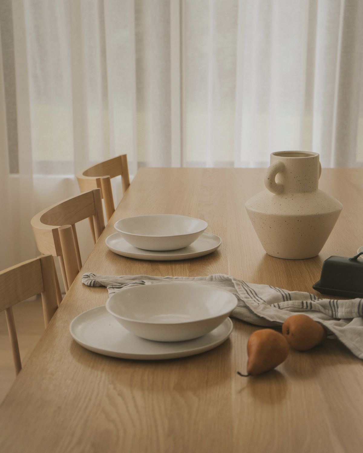 Tamminen ruokapöytä, valkoiset lautaset ja kulhot sekä keraaminen kannu.